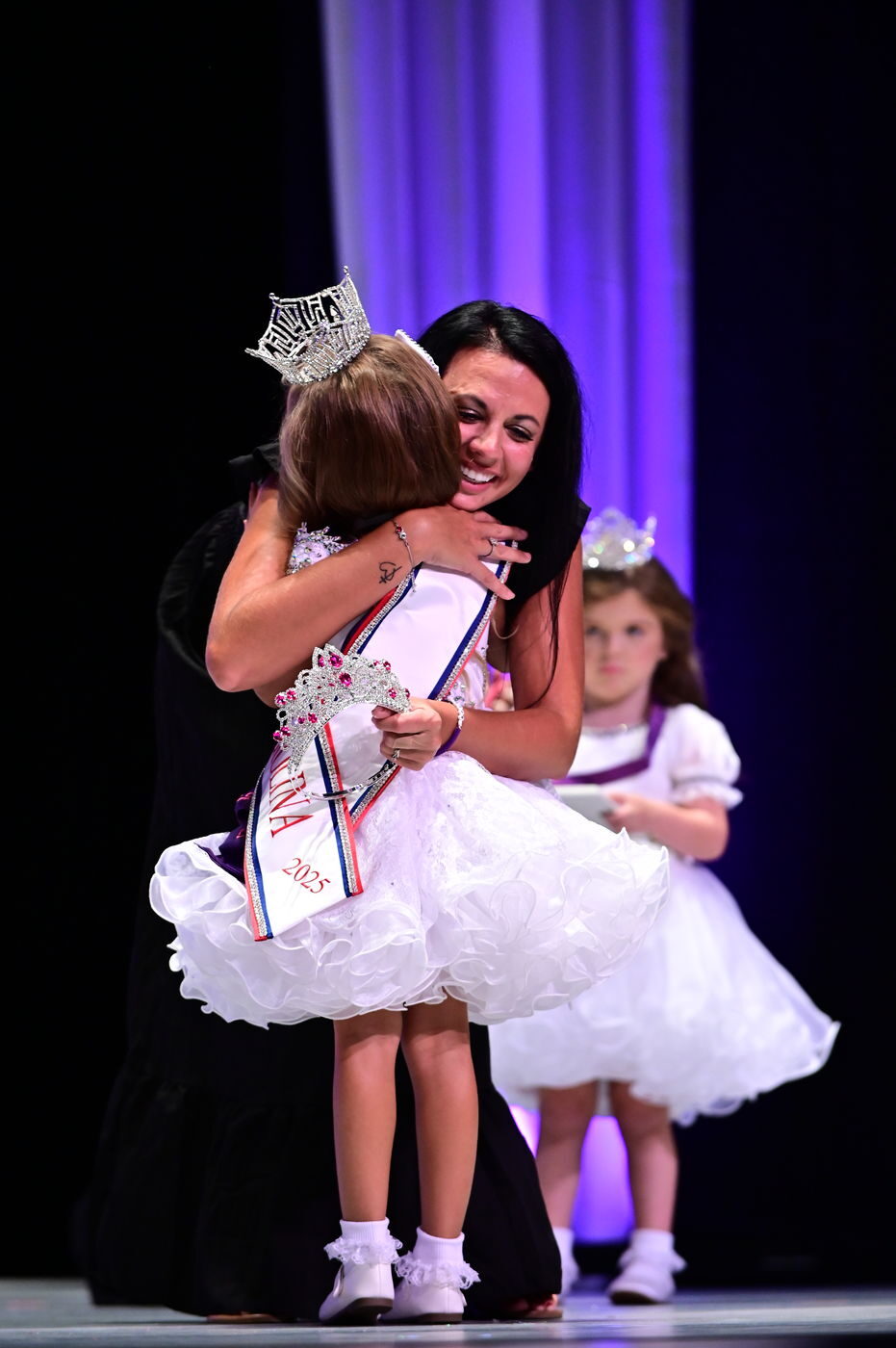 A crowning moment, a queen embracing a contestant while a small girl watches